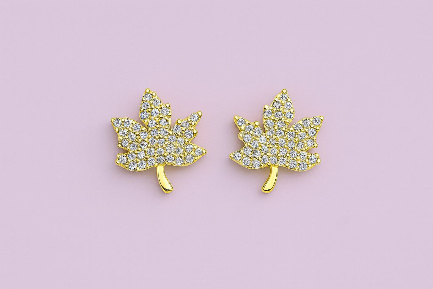 Golden maple leaf stud earrings with sparkling cubic zirconia stones on a pink background