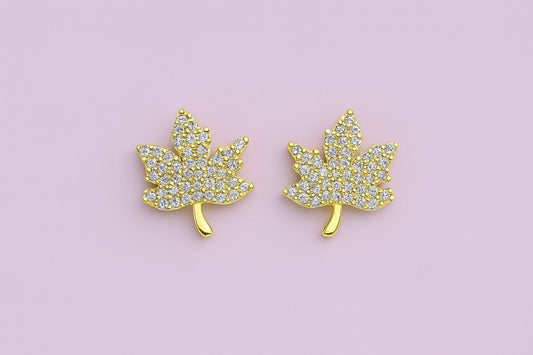 Golden maple leaf stud earrings with sparkling cubic zirconia stones on a pink background